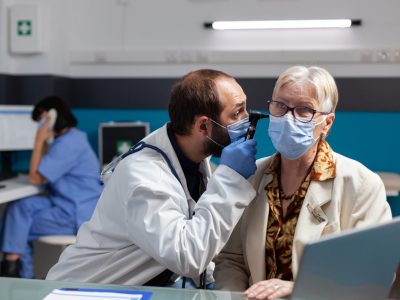 Physician consulting mature woman with otoscope at checkup visit, doing ear examination to treat infection during covid 19 pandemic. Doctor using otoscopy tool at consultation appointment.